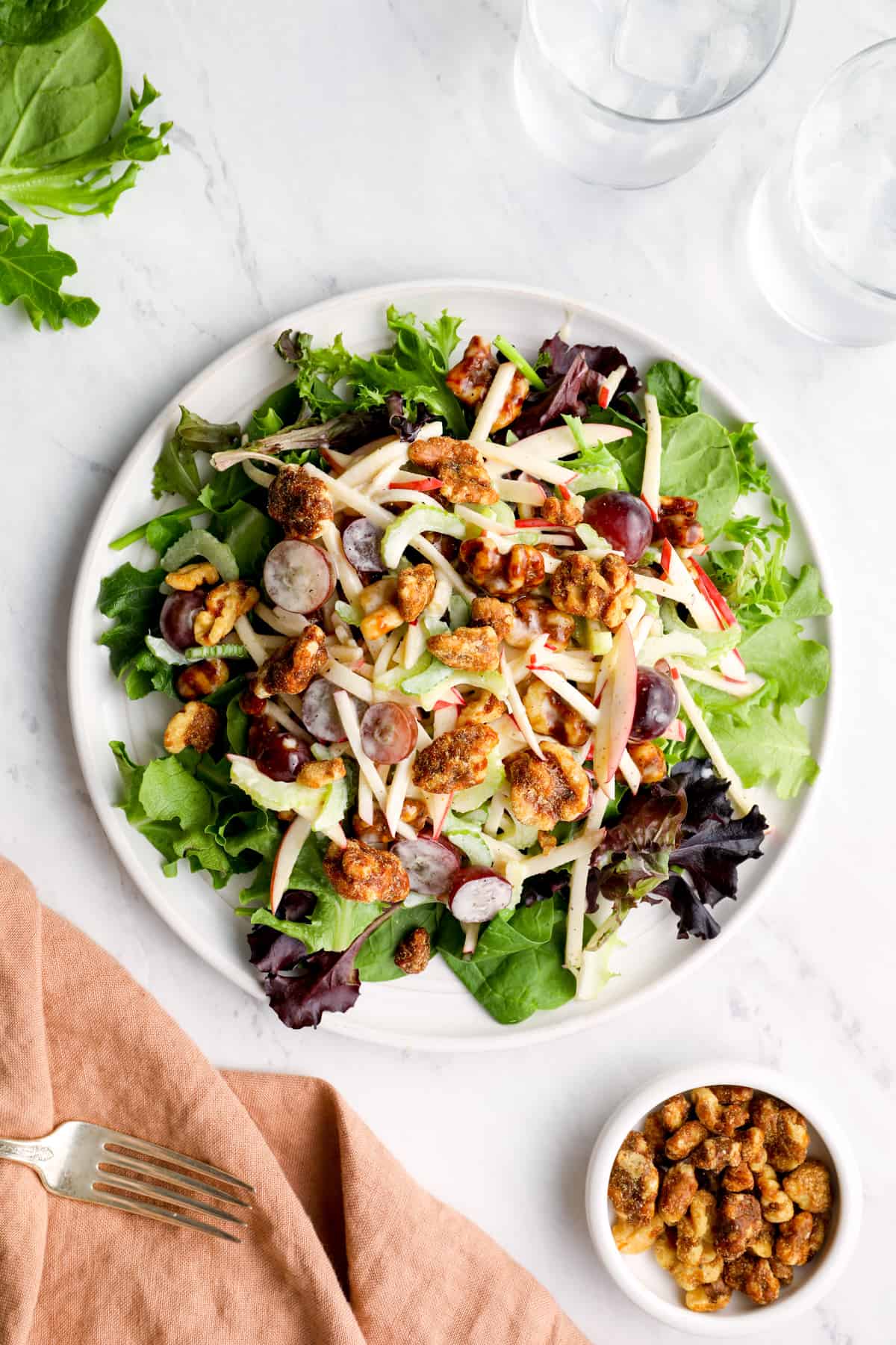 Waldorf salad on a table along with a cloth napkin, fork, glasses of water, and a small bowl of walnuts