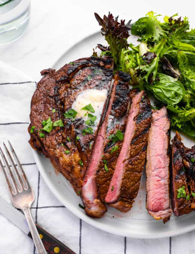 distant three-quarters view of a sliced marinated ribeye steak on a white plate with salad greens.