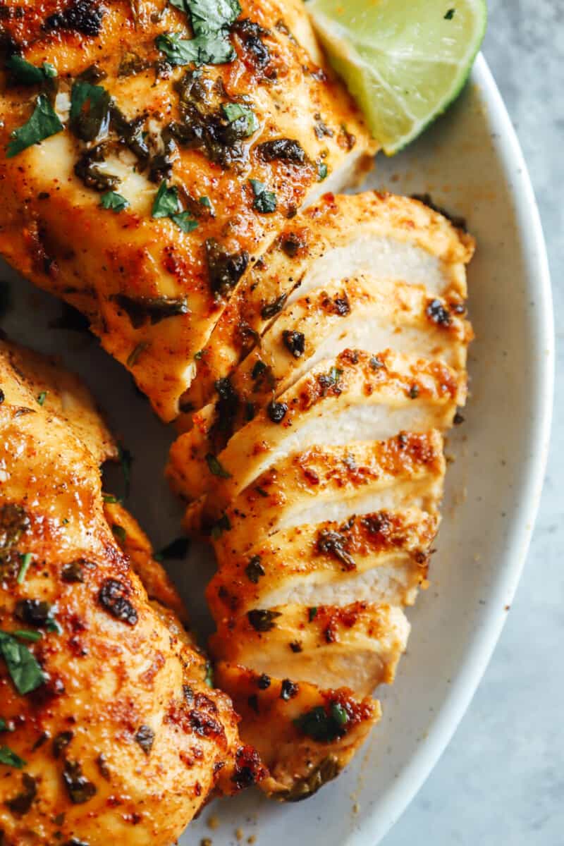 close up of a partially sliced cilantro lime chicken breast surrounded by more chicken breasts and lime wedges on a white plate.