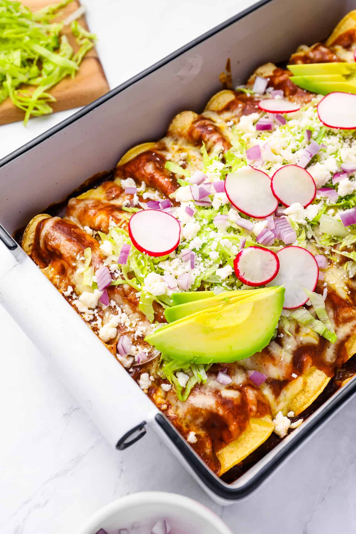 chicken enchiladas in a rectangular baking pan topped with lettuce, radish, onion, and avocado.