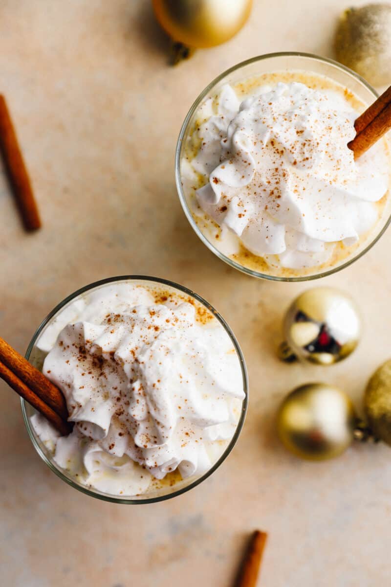 overhead view of 2 glasses of crockpot eggnog with whipped cream and cinnamon sticks.