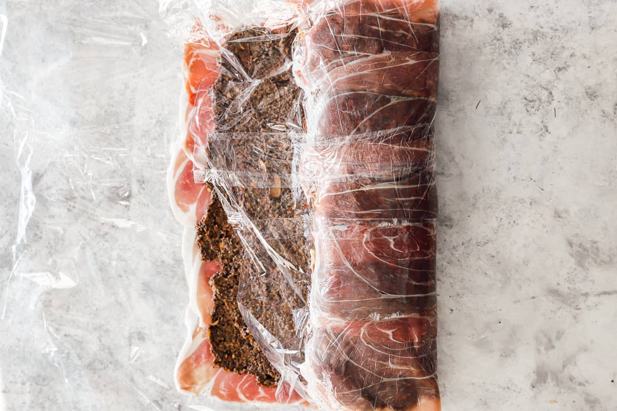 beef tenderloin covered in mushrooms and prosciutto being wrapped in plastic wrap.