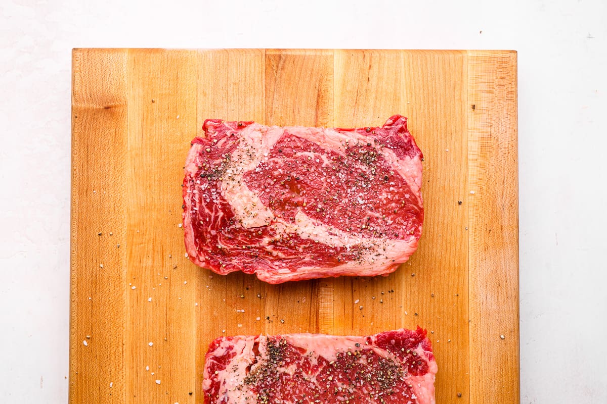 2 seasoned ribeye steaks on a wooden cutting board.