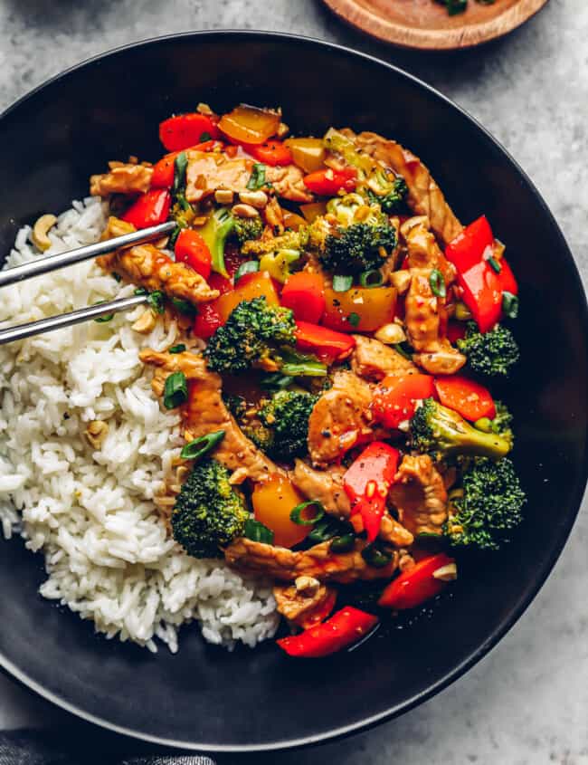 metal chopsticks grabbing a piece of pork from pork stir fry with white rice in a black bowl.