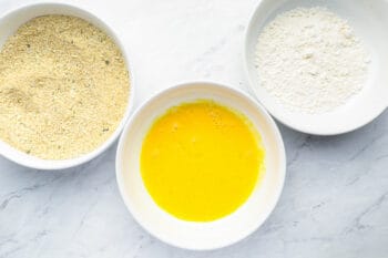 a dredging station consisting of seasoned breadcrumbs in a white bowl, scrambled raw eggs in a white bowl, and flour in a white bowl.