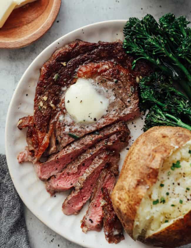 overhead view of a sliced pan seared ribeye on a white plate topped with butter and a side of baked potato and broccoli rabe.