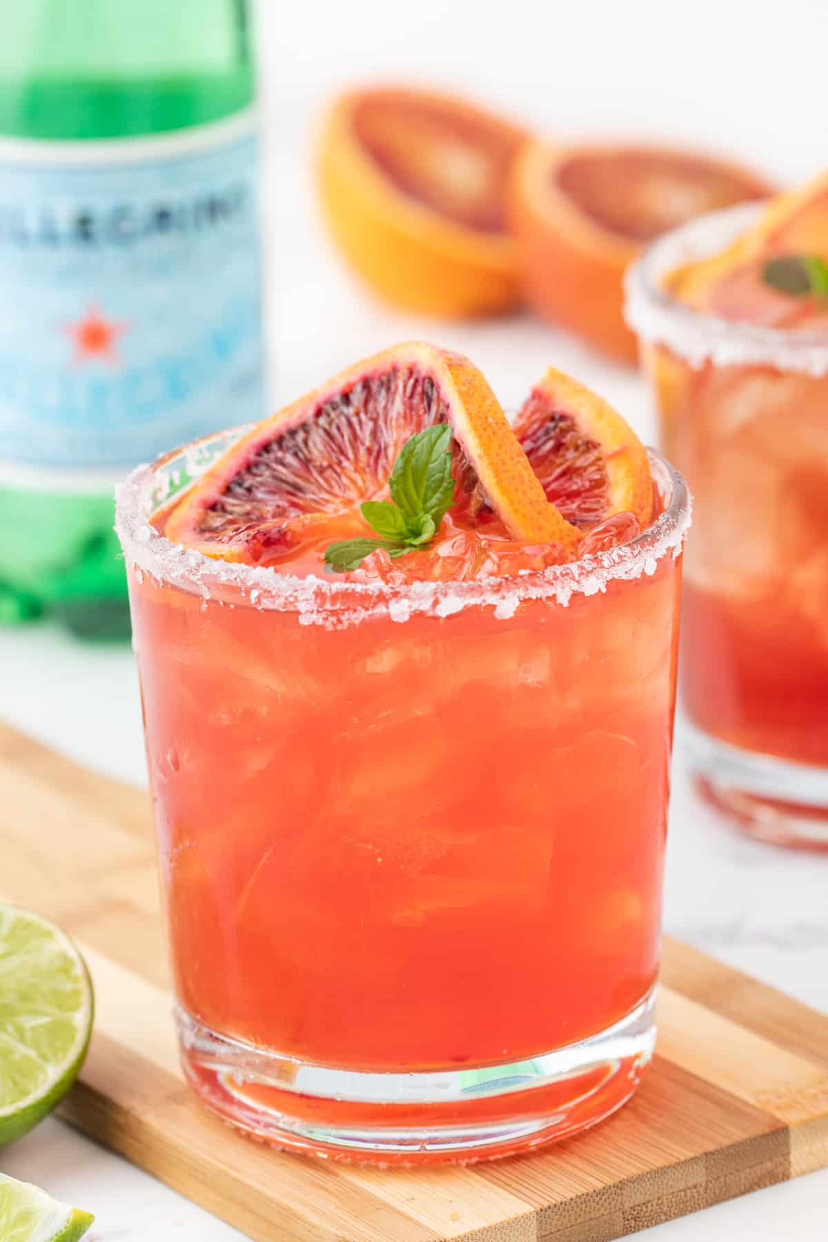 close up three-quarters view of a blood orange margarita on a wooden cutting board garnished with a blood orange slice and mint leaves.