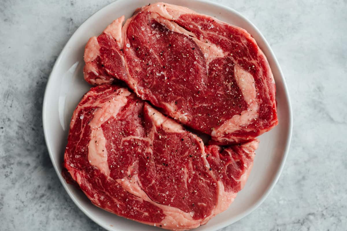 overhead view of 2 raw ribeye steaks on a white plate.