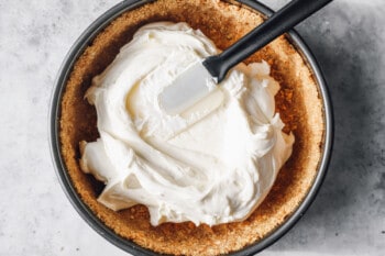 no bake cheesecake filling spread into a graham cracker crust in a springform pan with a rubber spatula.