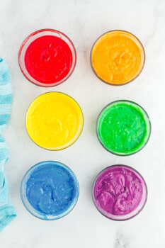 overhead view of cake batter in 6 glass bowls, colored red, orange, yellow, green, blue, and purple.