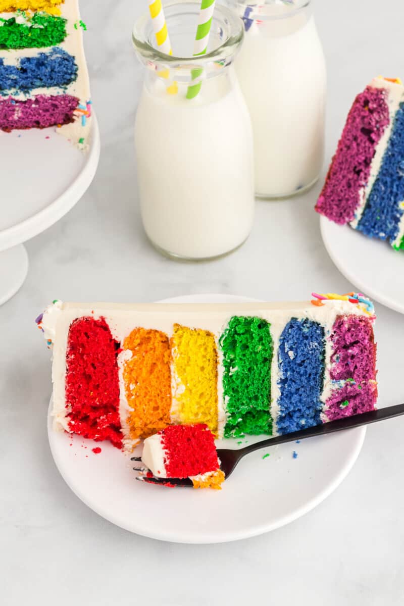 a slice of rainbow cake on its side on a white plate with a fork.