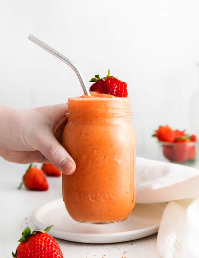 a hand grabbing a strawberry mango smoothie in a mason jar with a stainless steel straw.