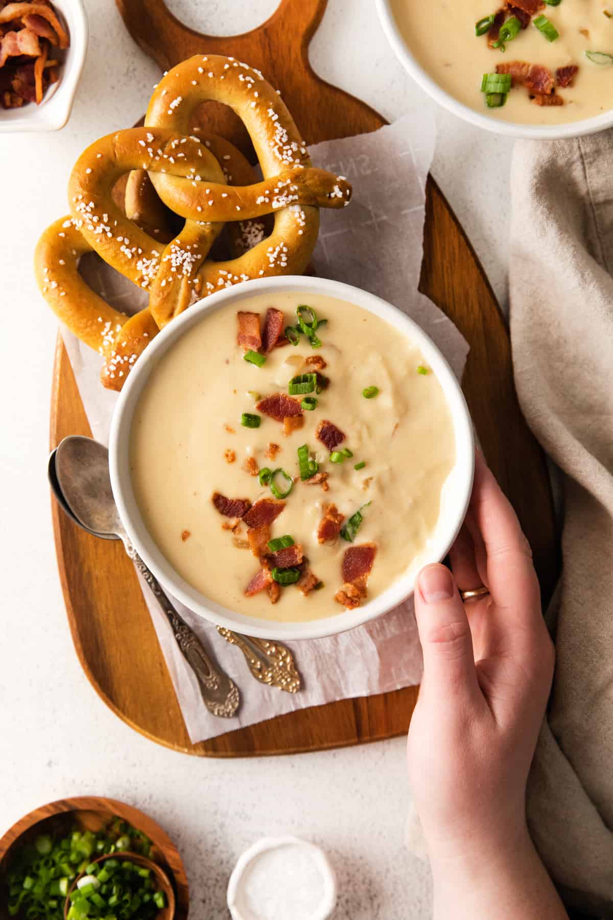 overhead view of a hand grabbing a serving of beer cheese soup topped with bacon in a white bowl from a cutting board with 2 soft pretzels.