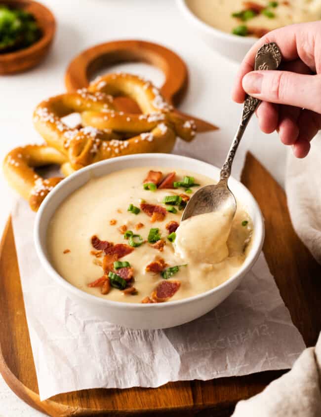 three-quarters view of a hand dipping a spoon into a serving of beer cheese soup in a white bowl on a cutting board with 2 soft pretzels.