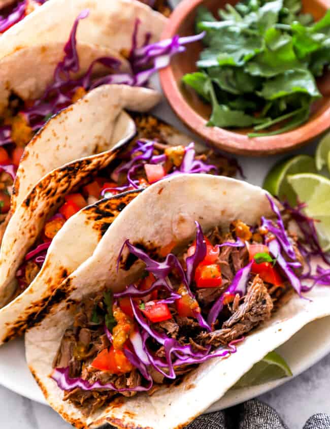 overhead view of 4 barbacoa tacos on a white plate with a bowl of cilantro.