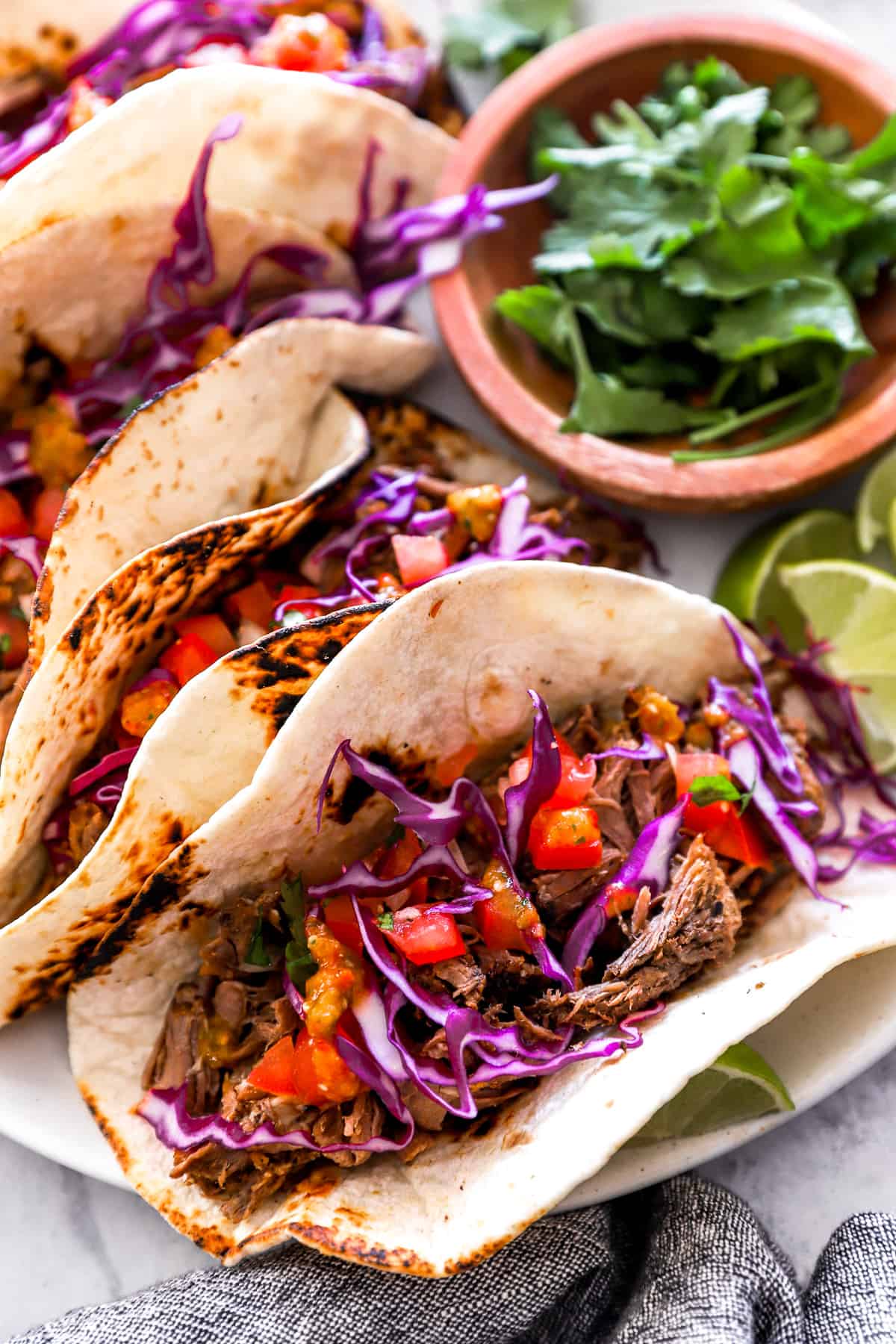 overhead view of 4 barbacoa tacos on a white plate with a bowl of cilantro.