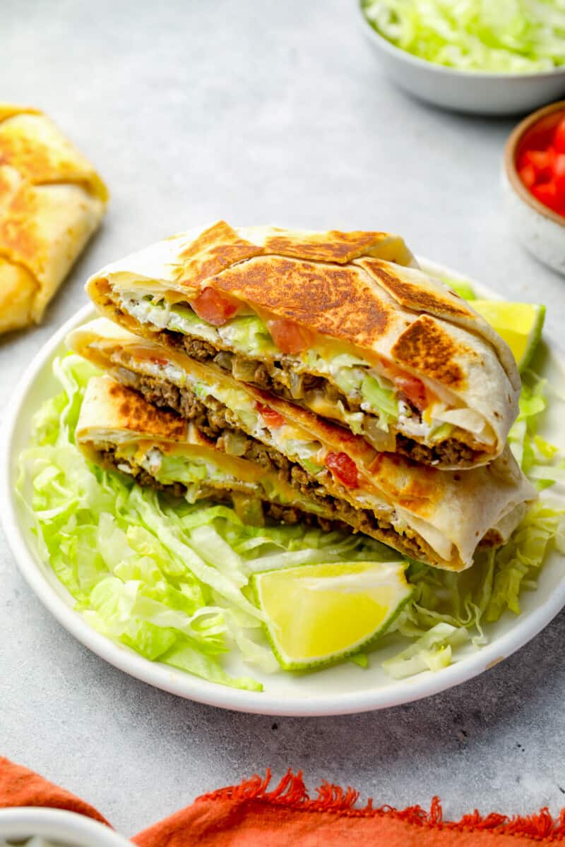 three-quarters view of a halved crunchwrap supreme stacked on top of itself on a white plate with lettuce and lime wedges.