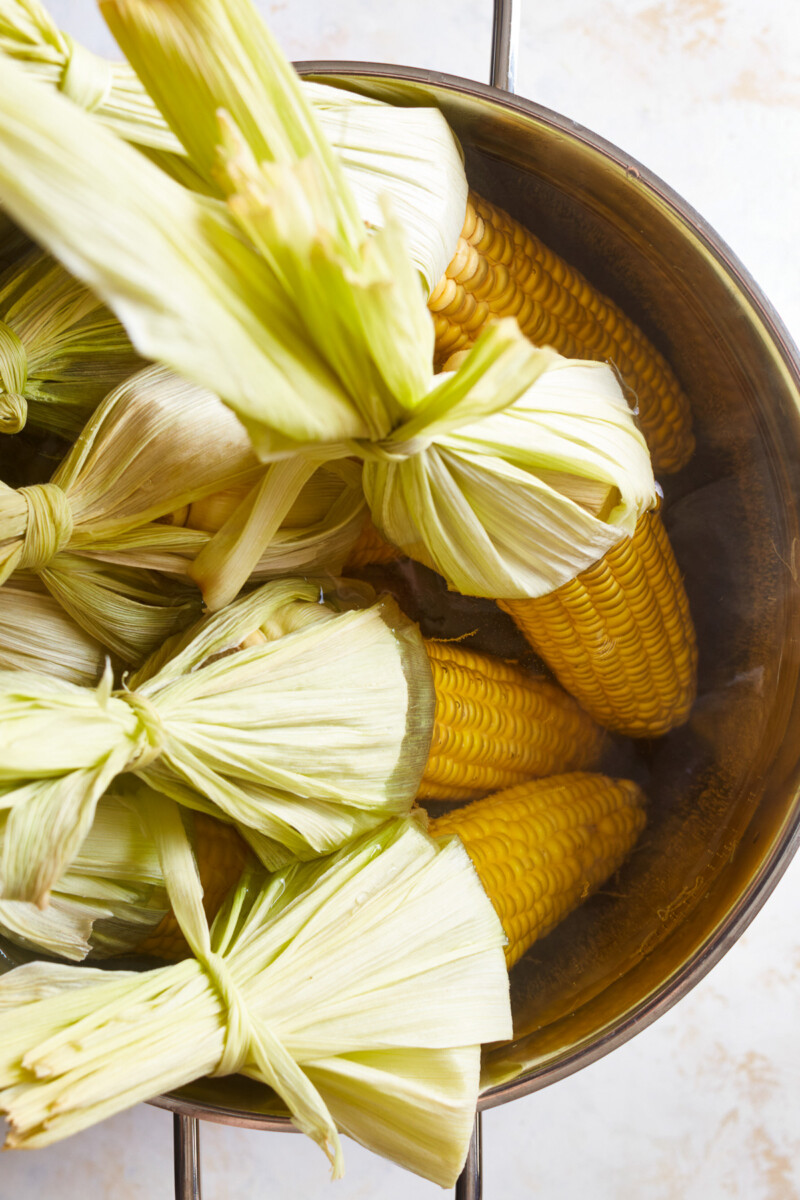 8 ears of corn pointed down into a deep pot of water with the husks pulled back and sticking up.