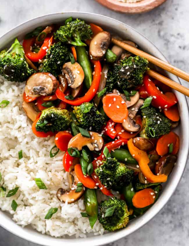 partial overhead view of vegetable stir fry in a white bowl with white rice and chopsticks.