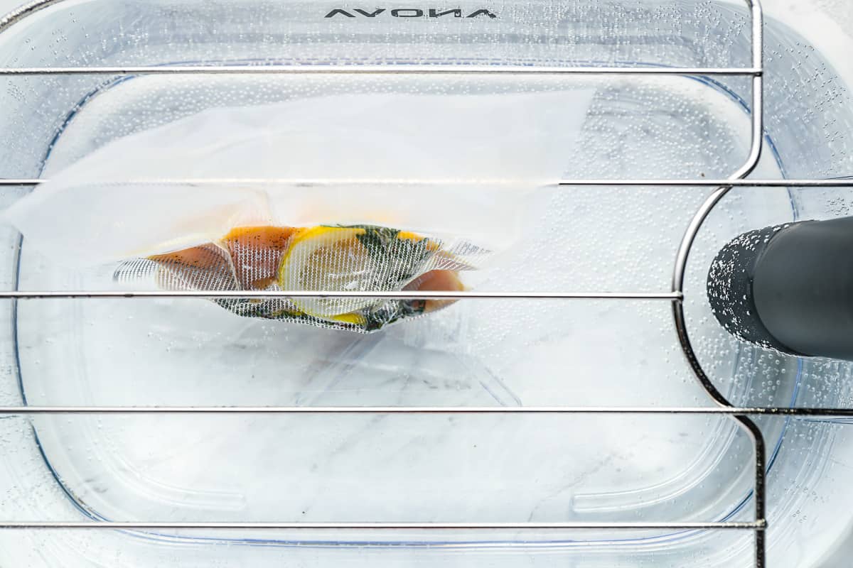 overhead view of a vacuum sealed bag filled with 2 raw chicken breasts, lemon slices, and herbs in a sous vide bath.