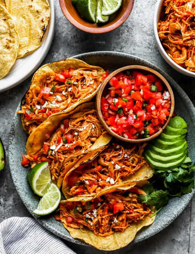 overhead view of 4 instant pot chicken tacos on a gray plate with salsa, avocados, and lime wedges.