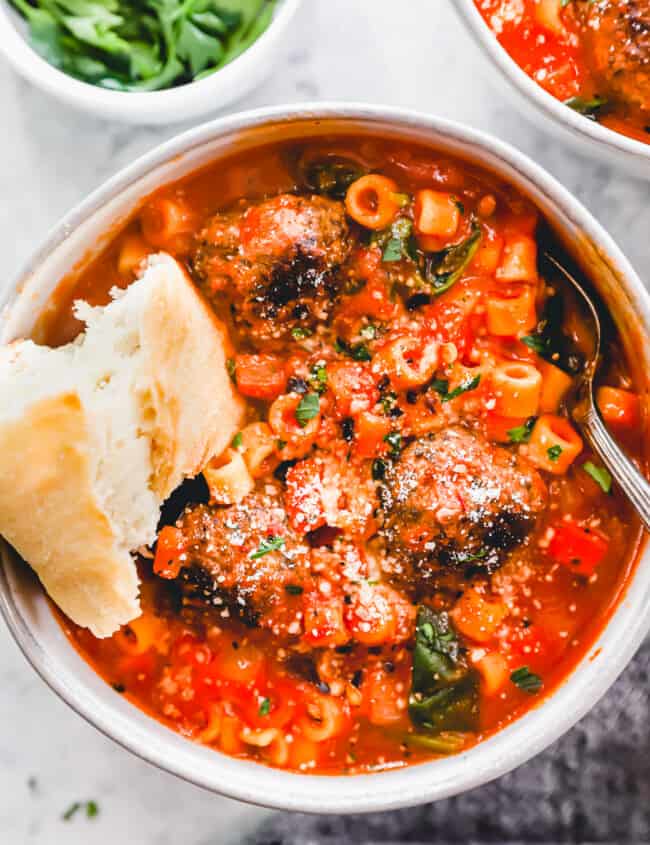 overhead view of a serving of meatball soup in a white bowl with a spoon and a piece of bread.