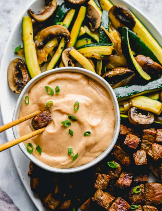 overhead view of a pair of chopsticks dipping a cube of steak into yum yum sauce in a white bowl.