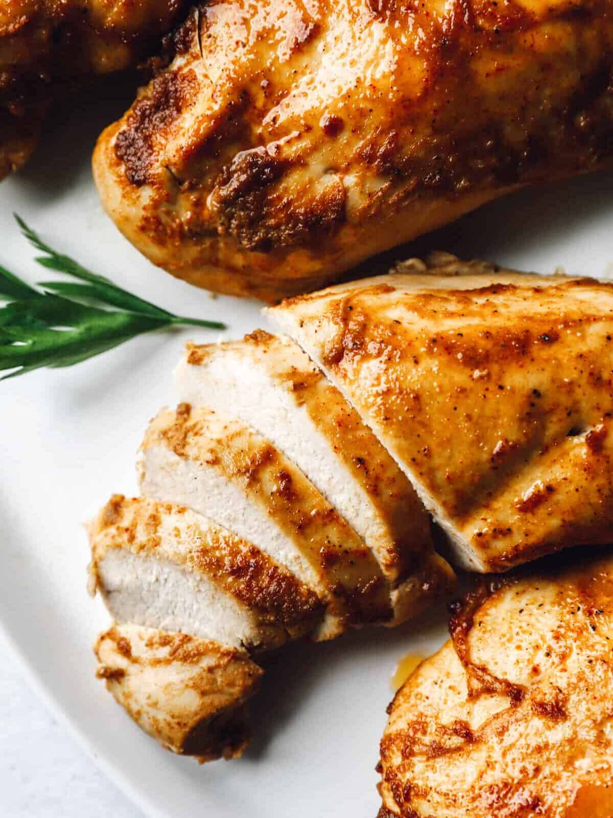 1 partially sliced chicken breast on a white platter next to 3 baked marinated chicken breasts.