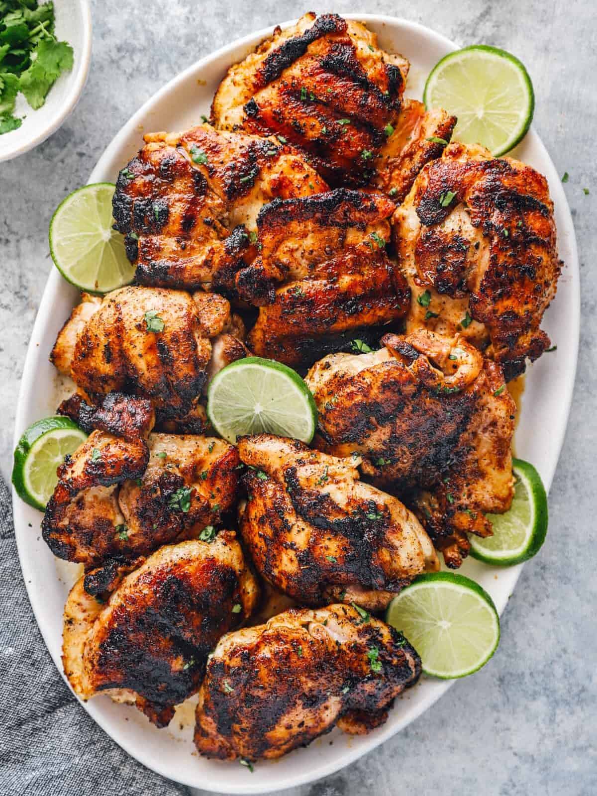 overhead view of seasoned grilled chicken thighs on a white oval serving platter with lime slices.