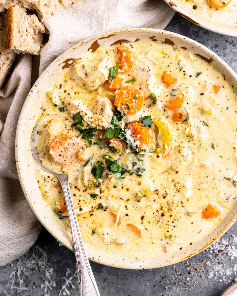 overhead view of creamy chicken soup in a bowl, slices of carrots, shredded chicken, and seasonings are visible on the surface, a soup spoon lays at the left-side of the bowl