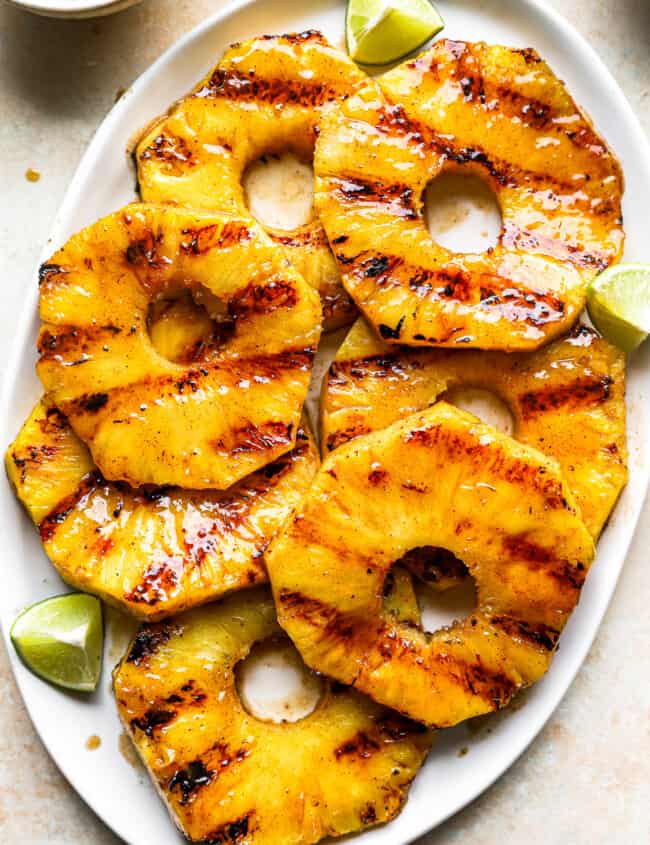 overhead view of slices of grilled pineapple on a white oval serving platter with lime wedges.