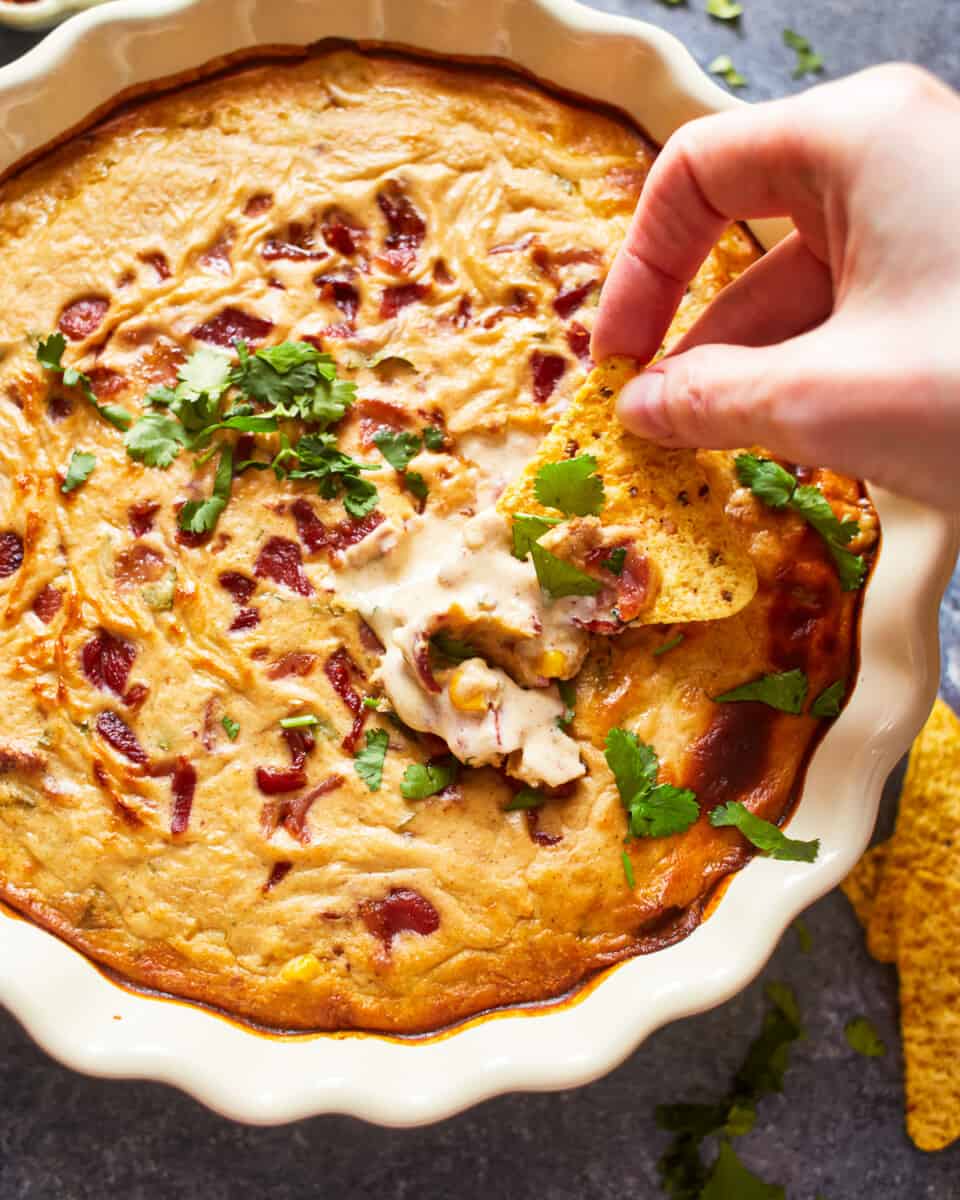 overhead view of a hand dipping a tortilla chip into hot corn and bacon cheese dip in a white round dish.