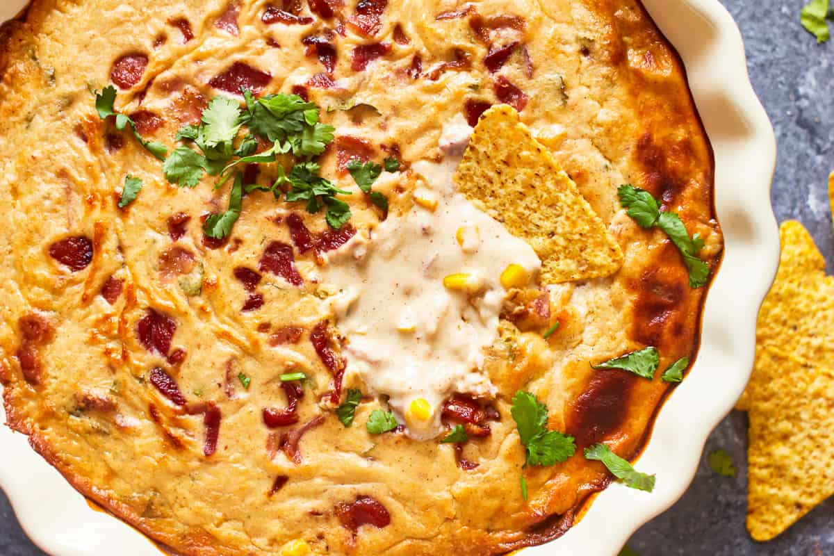 close up overhead view of a tortilla chip resting on top of hot corn and bacon cheese dip in a white round dish.