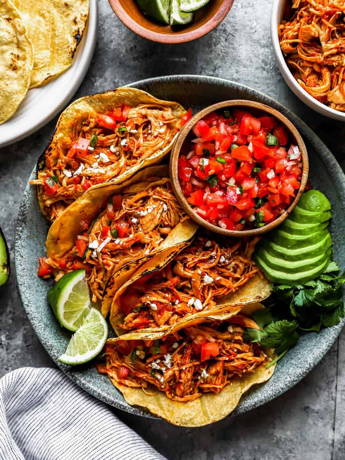 overhead view of 4 instant pot chicken tacos on a gray plate with salsa, avocados, and lime wedges.