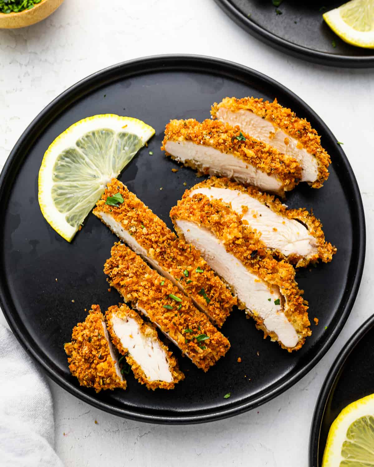 a sliced panko breaded chicken breast on a black plate with lemon wedges.