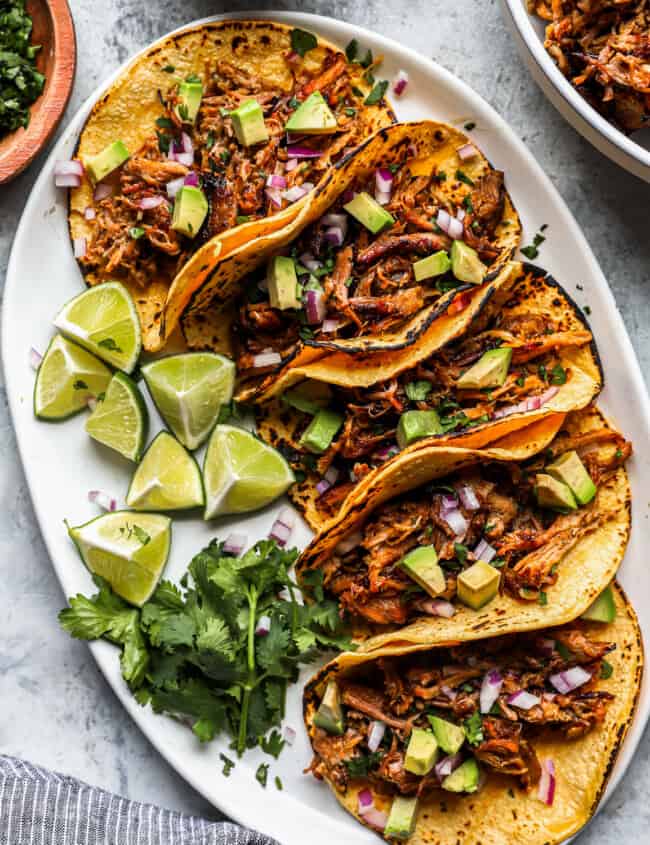 overhead view of 5 carnitas tacos on a white oval serving platter with limes.