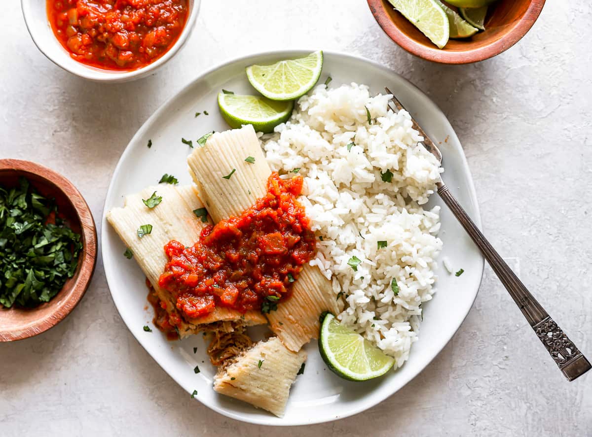 overhead view of 2 homemade corn tamales on a white plate with cilantro rice, salsa, and a fork, one tamale has been sliced into.