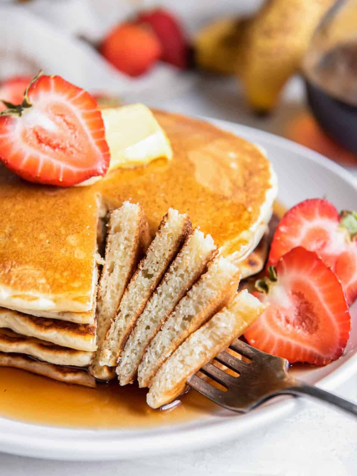 a forkful of buttermilk pancakes resting on a plate of buttermilk pancakes with syrup, butter, and a halved strawberry.