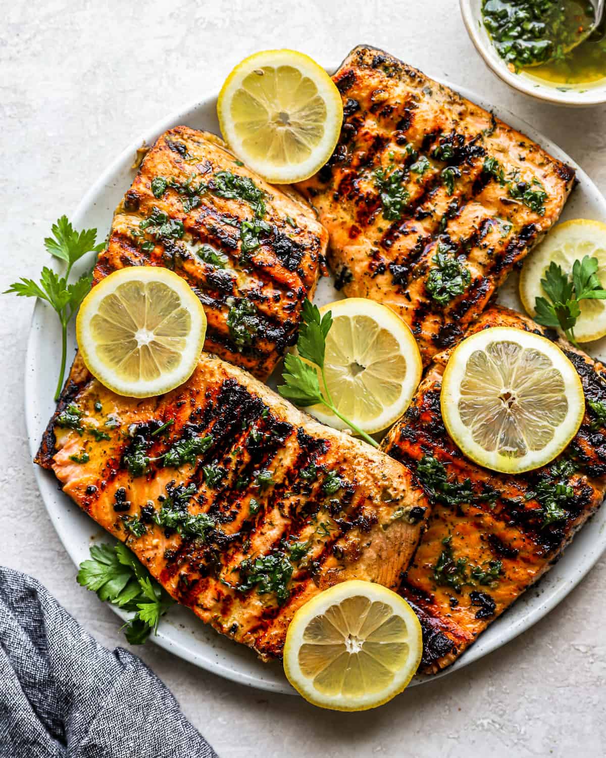 overhead view of 4 overlapping grilled salmon fillets on a white plate topped with herbs and lemon slices.