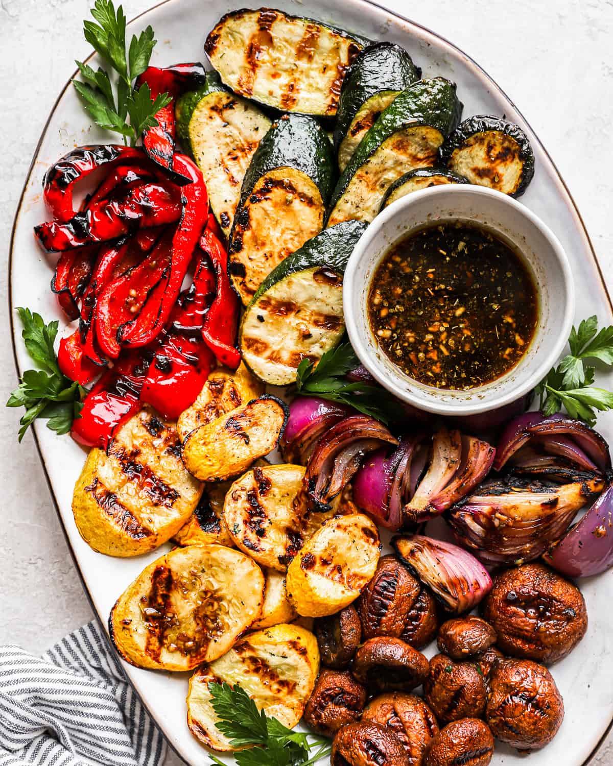 overhead view of grilled vegetables on a white oval plate with marinade in a small bowl.