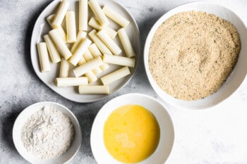 halved mozzarella sticks on a white plate next to bowls filled with flour, eggs, and breadcrumbs.