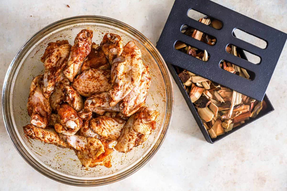 seasoned raw chicken wings in a glass bowl next to a smoker box filled with wood chips.