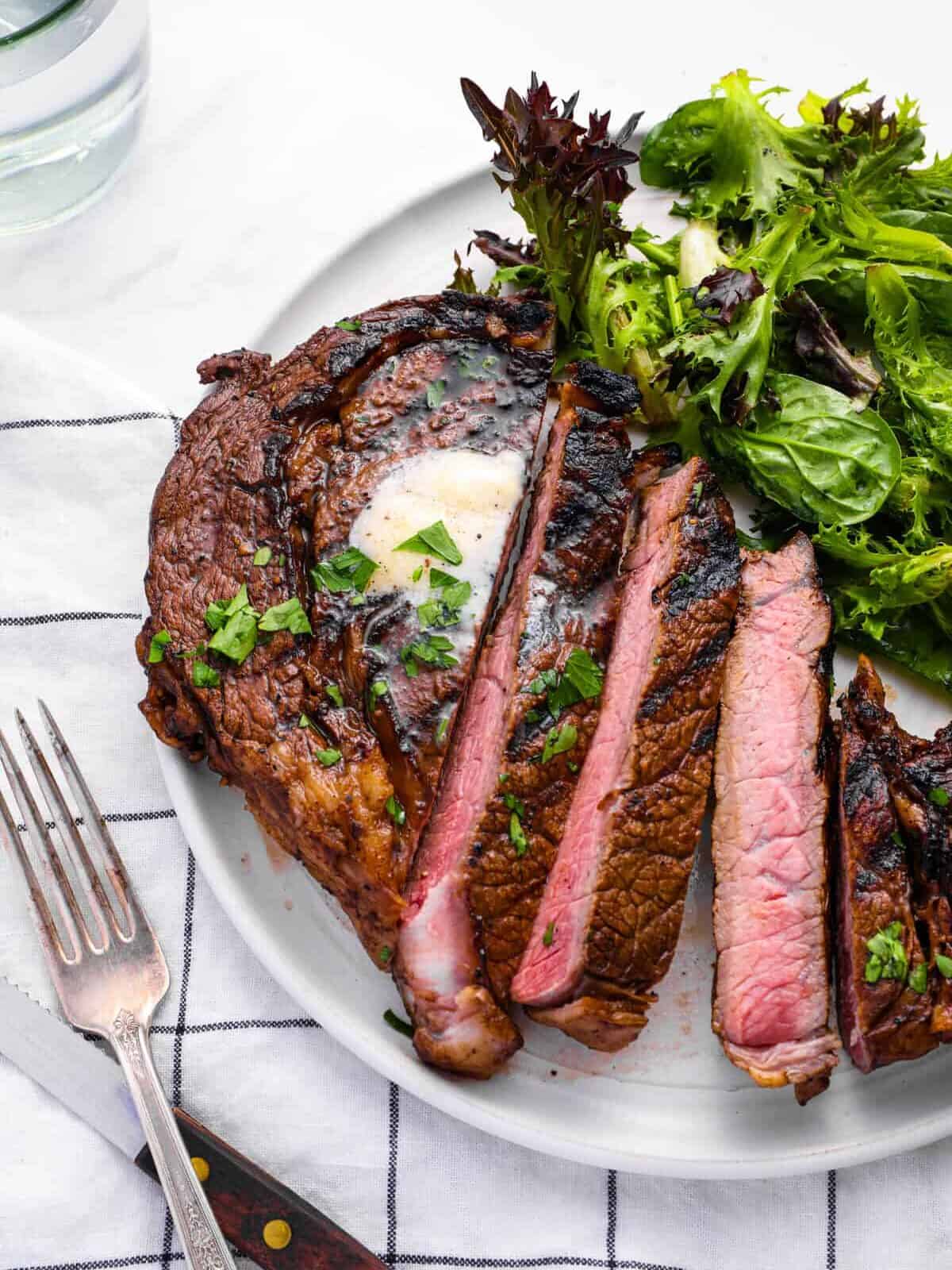 distant three-quarters view of a sliced marinated ribeye steak on a white plate with salad greens.