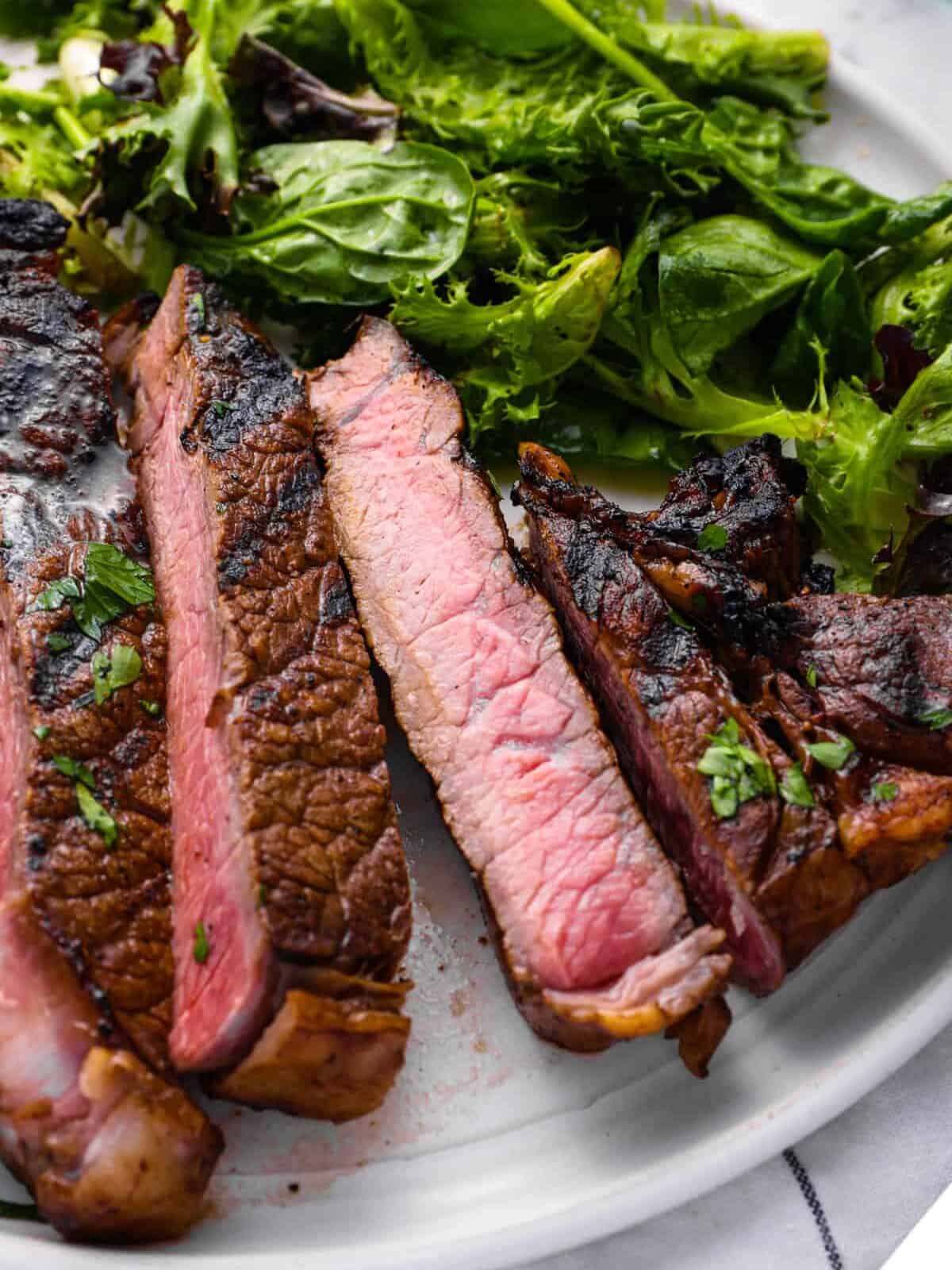 close up view of a sliced marinated ribeye steak on a white plate with salad greens.