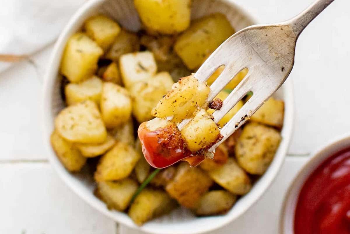 close-up overhead view of a forkful of breakfast potatoes hovering over a serving of breakfast potatoes in a white bowl.