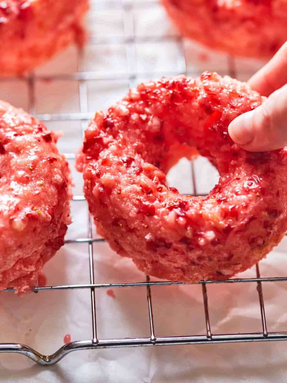 close up three-quarters view of a hand lifting a strawberry donut from a wire cooling rack.