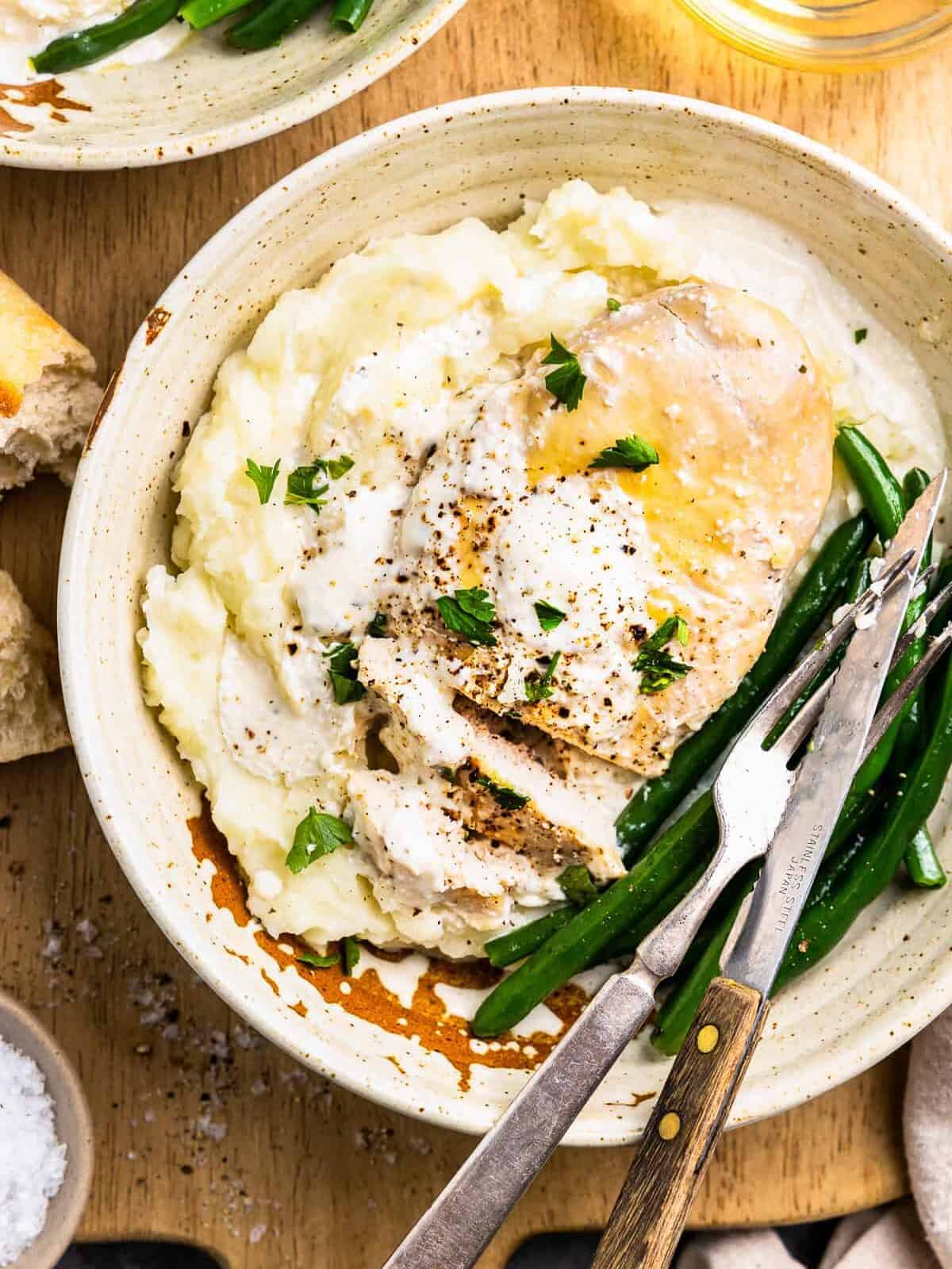 close-up overhead view of crockpot ranch chicken on a white plate with green beans, mashed potatoes, and a fork and knife.