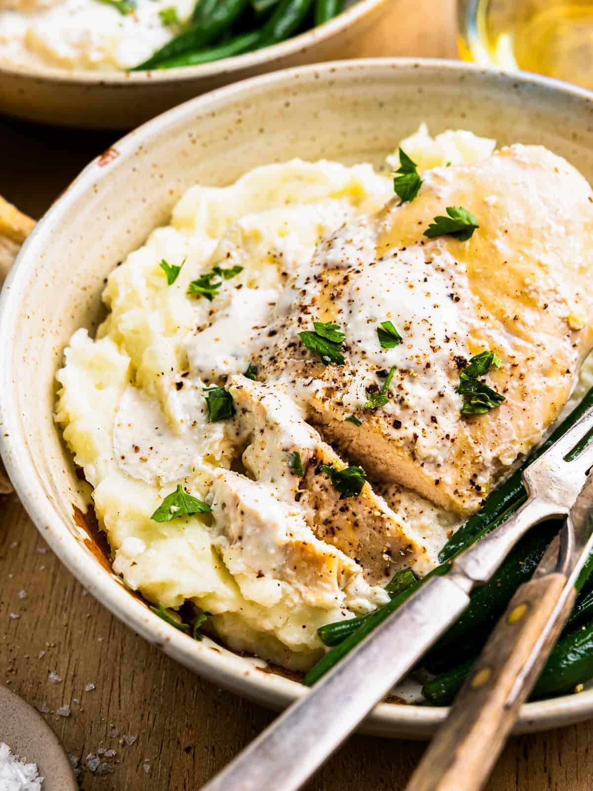 three-quarters view of crockpot ranch chicken on a white plate with green beans, mashed potatoes, and a fork and knife.