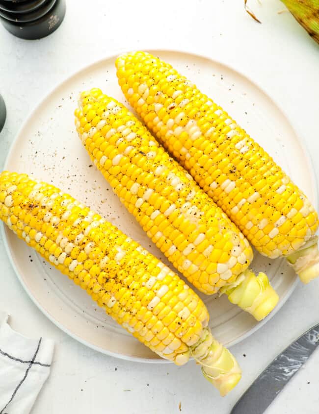 overhead view of 3 grilled corn cobs on a white plate seasoned with salt and pepper.