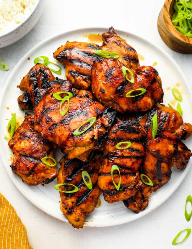 overhead view of grilled huli huli chicken thighs on a white plate topped with green onions.